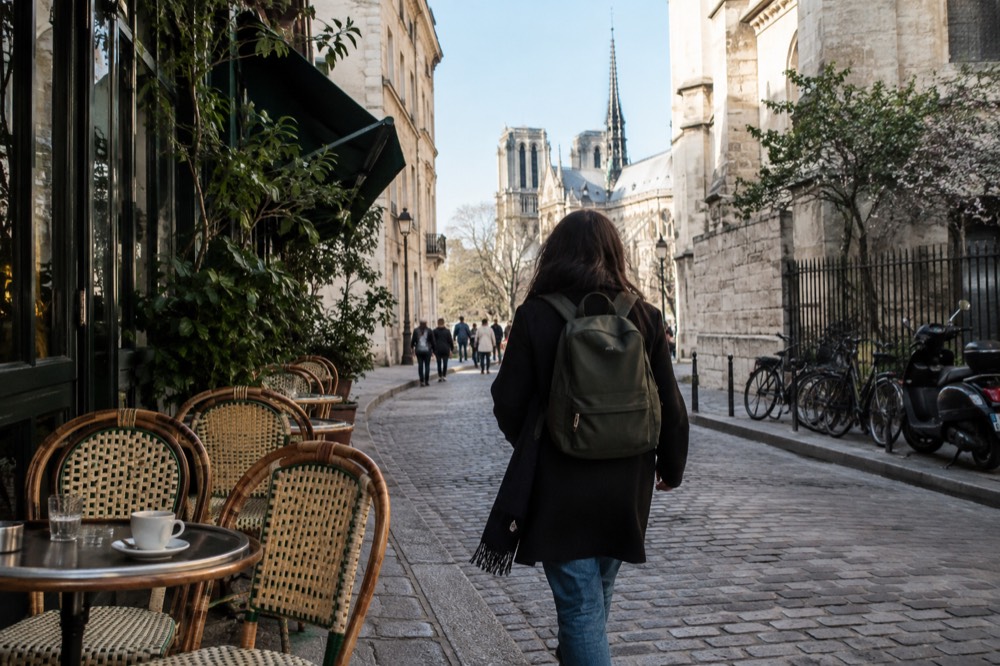 Un voyageur avec un petit sac à dos passant devant des terrasses de café dans une rue de Paris par un après-midi clair