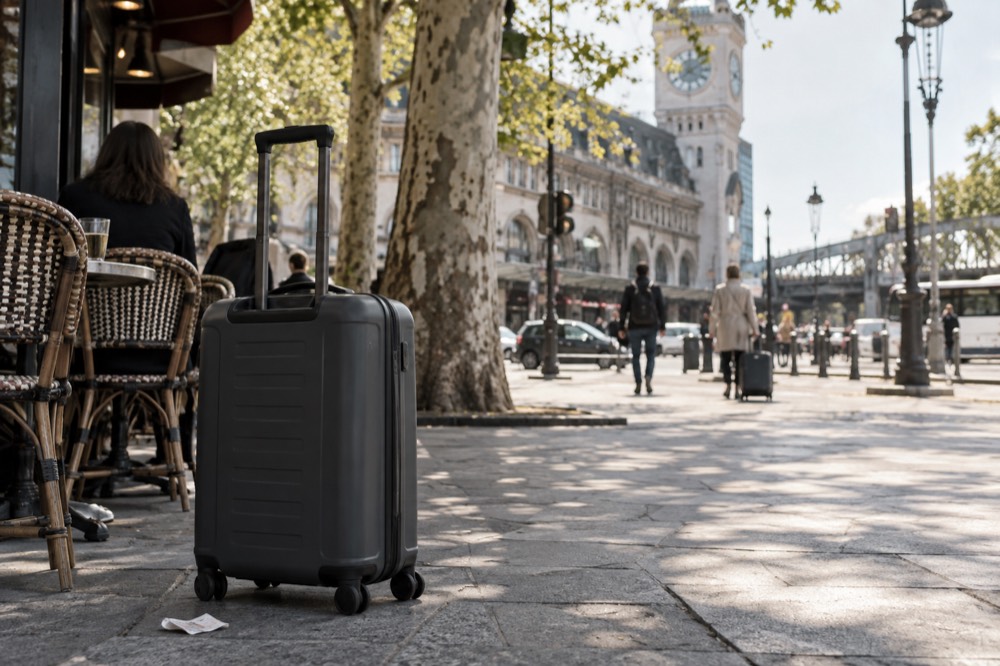Une valise sur un large trottoir parisien ensoleillé, devant la terrasse d'un café d'angle