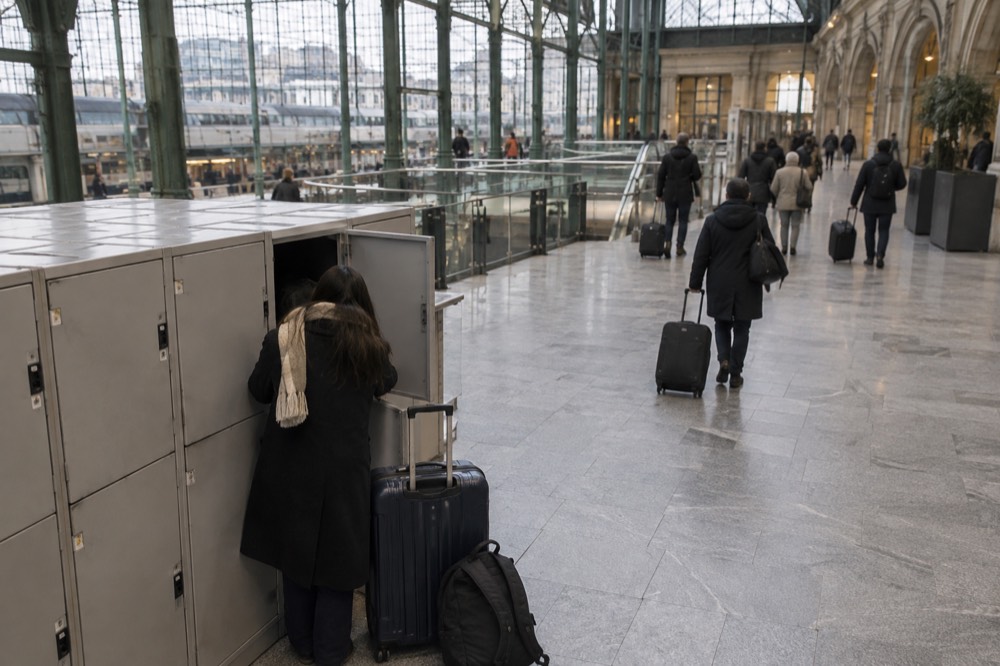 Hall d'une gare avec un espace de casiers de consigne et des voyageurs passant avec des chariots