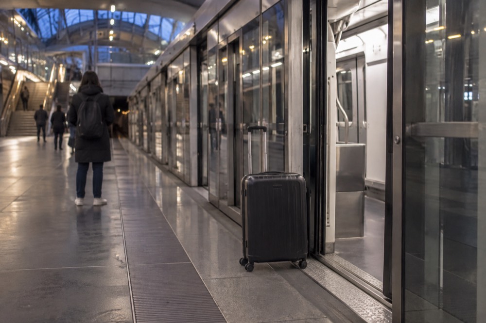 Un quai de métro moderne avec une porte d'ascenseur ouverte et une valise à roulettes à côté d'un voyageur qui attend une rame sans conducteur