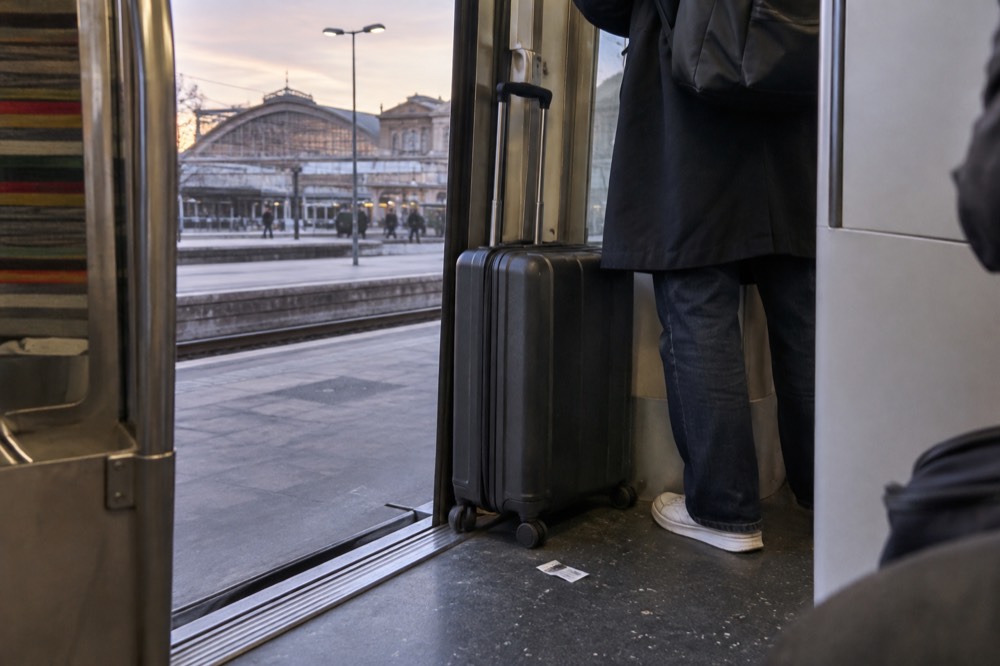 L'intérieur d'un train de banlieue avec une valise à roulettes maintenue en place dans le renfoncement de la porte, à côté d'un voyageur debout