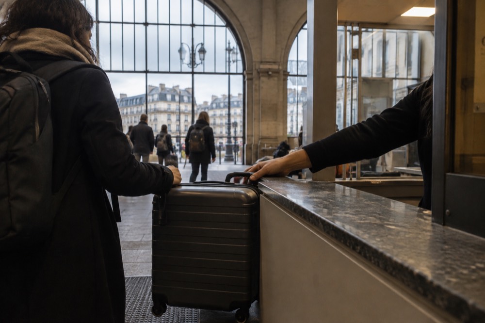 A traveller handing a tagged suitcase across a shop counter in Paris
