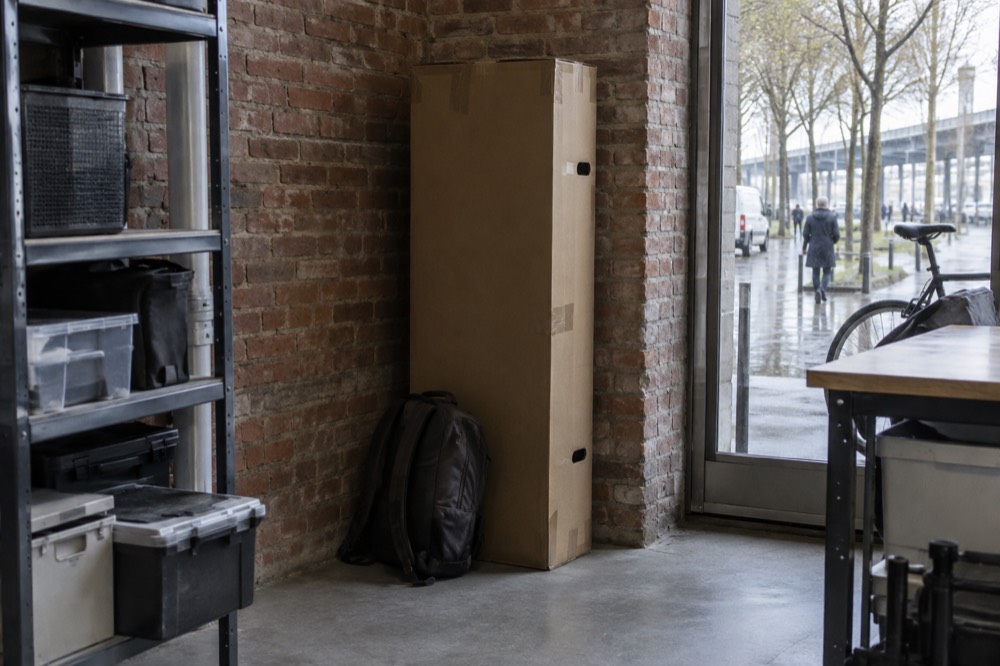 A cardboard bike box standing upright in the corner of a shop storage space